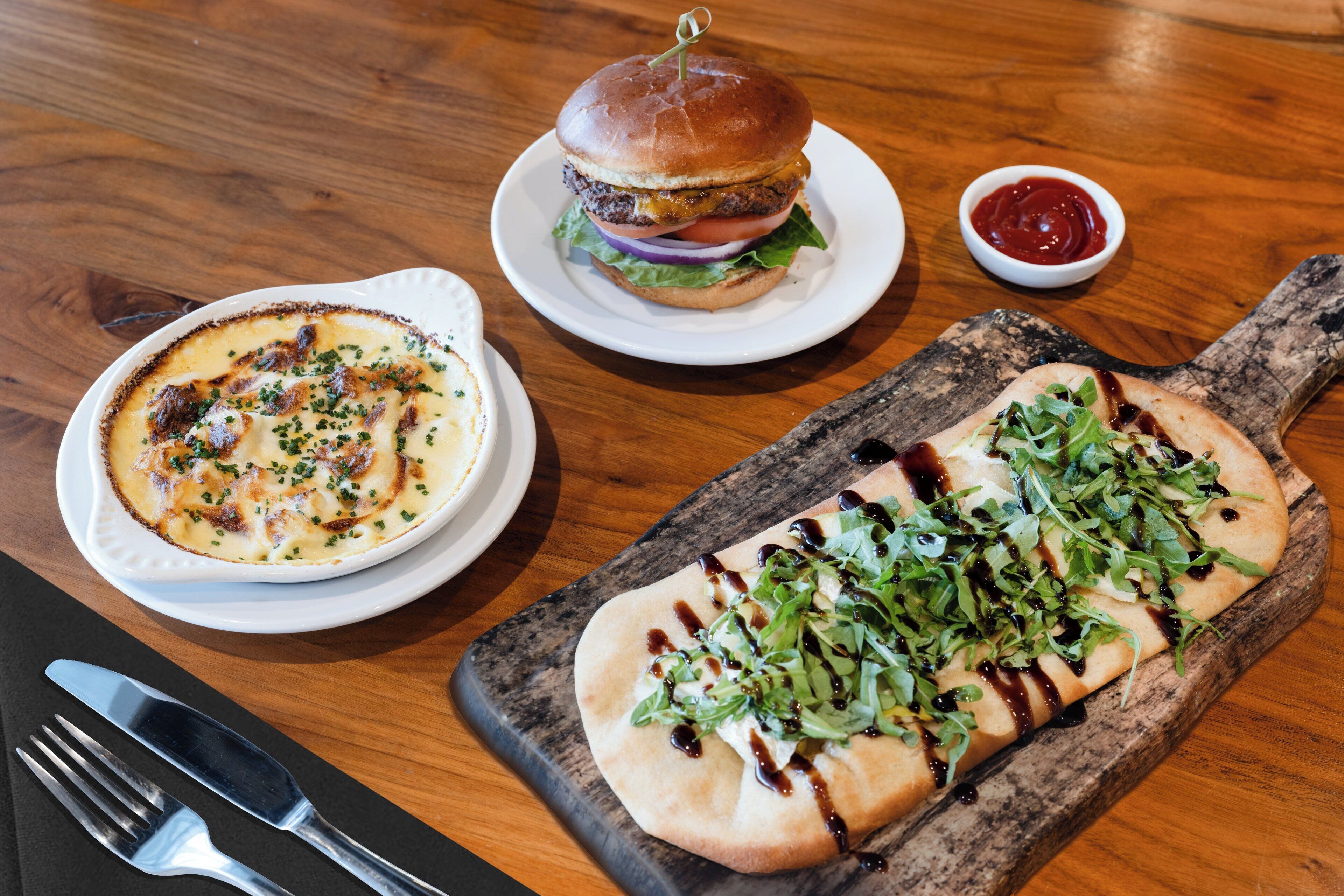A table with a burger, a bowl of soup, and a pizza on a wooden board.