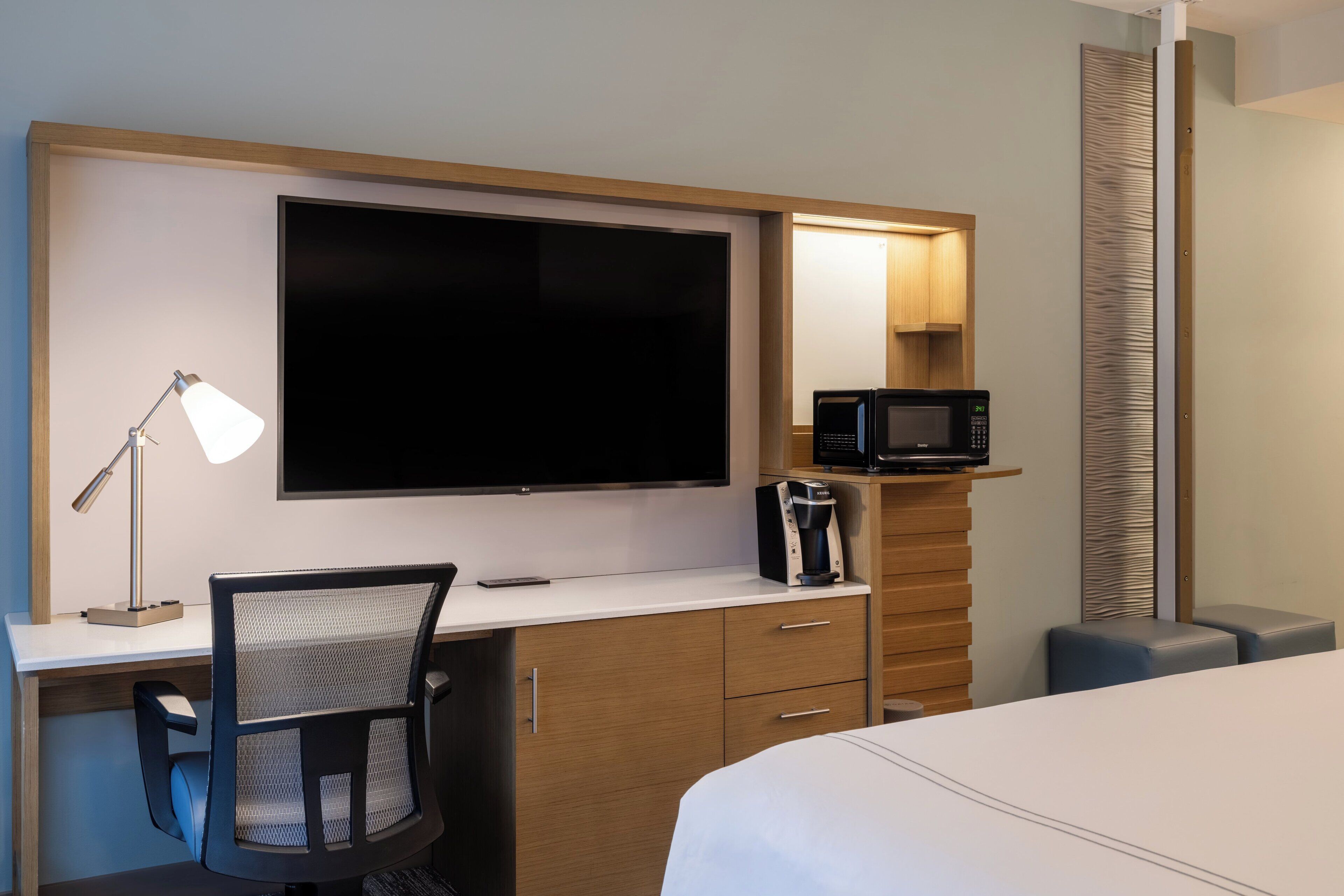 Ein Hotelzimmer mit einem großen Flachbildfernseher, einem Schreibtisch mit Lampe, einer Mikrowelle, einer Kaffeemaschine, einem Sessel und einem Bett.