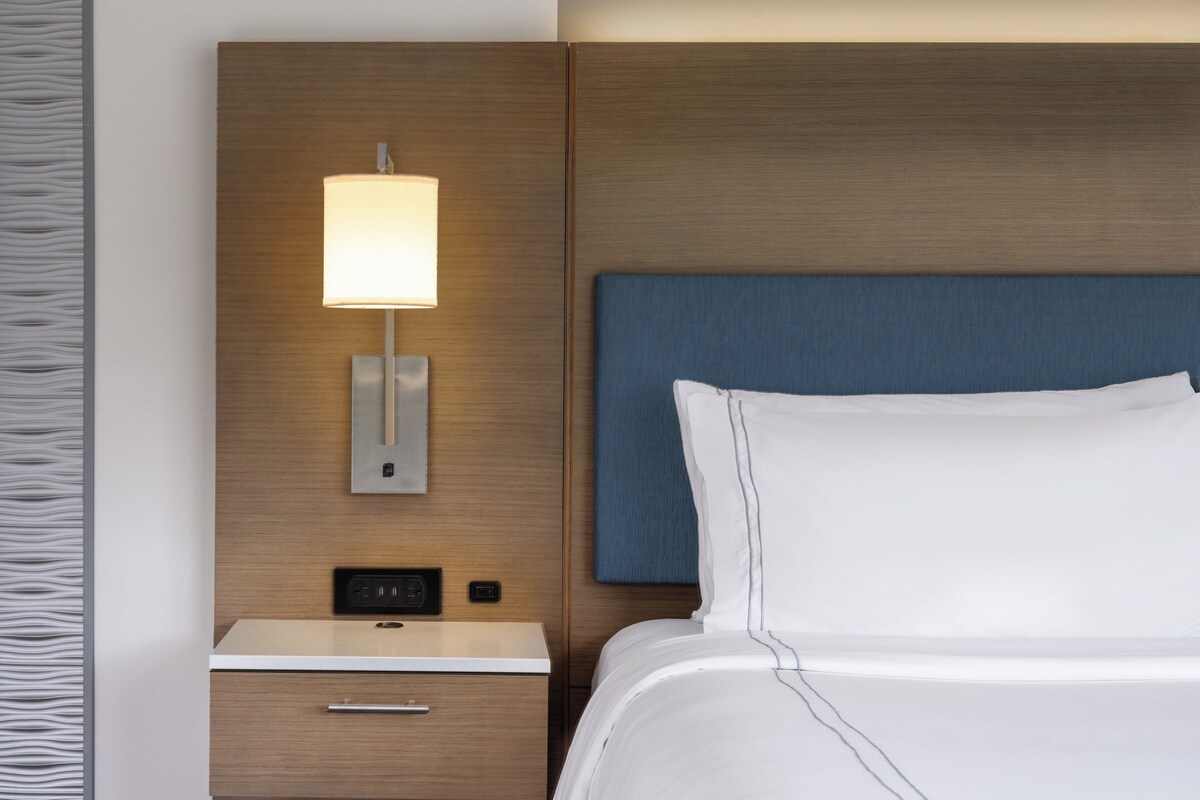 Een hotelkamer met een houten hoofdbord, een nachtkastje met een lamp en een bed met witte lakens.