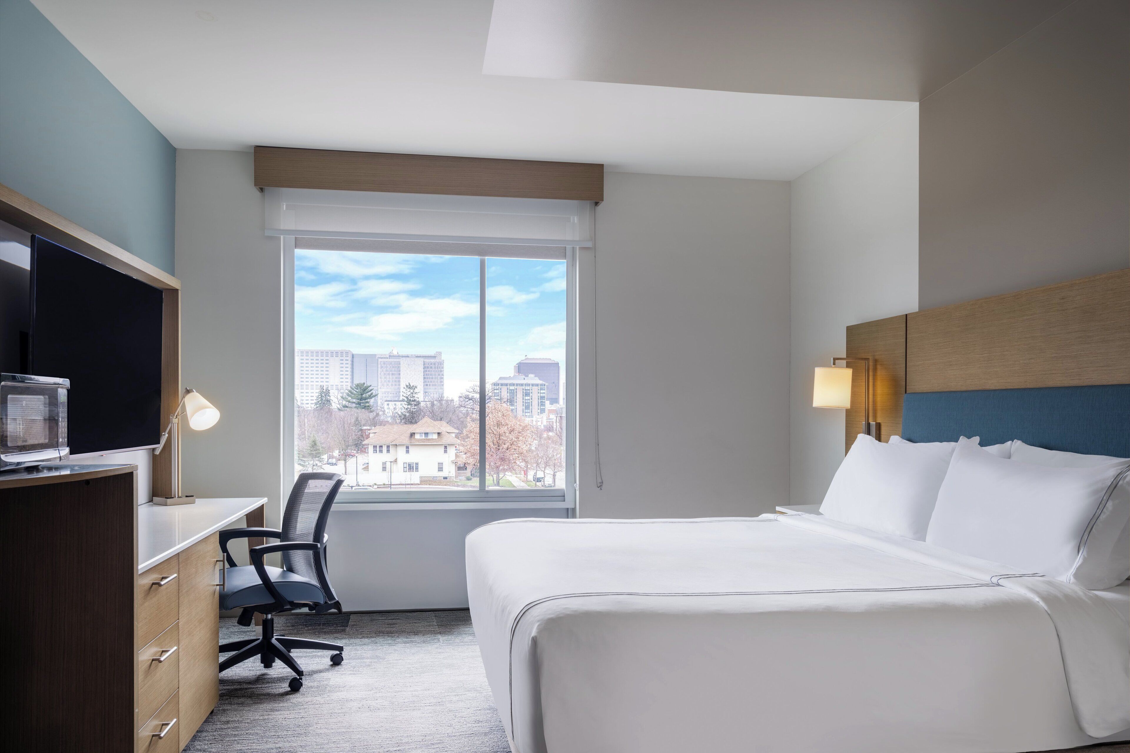 A hotel room with a bed, desk, chair, TV, and a window with a city view.