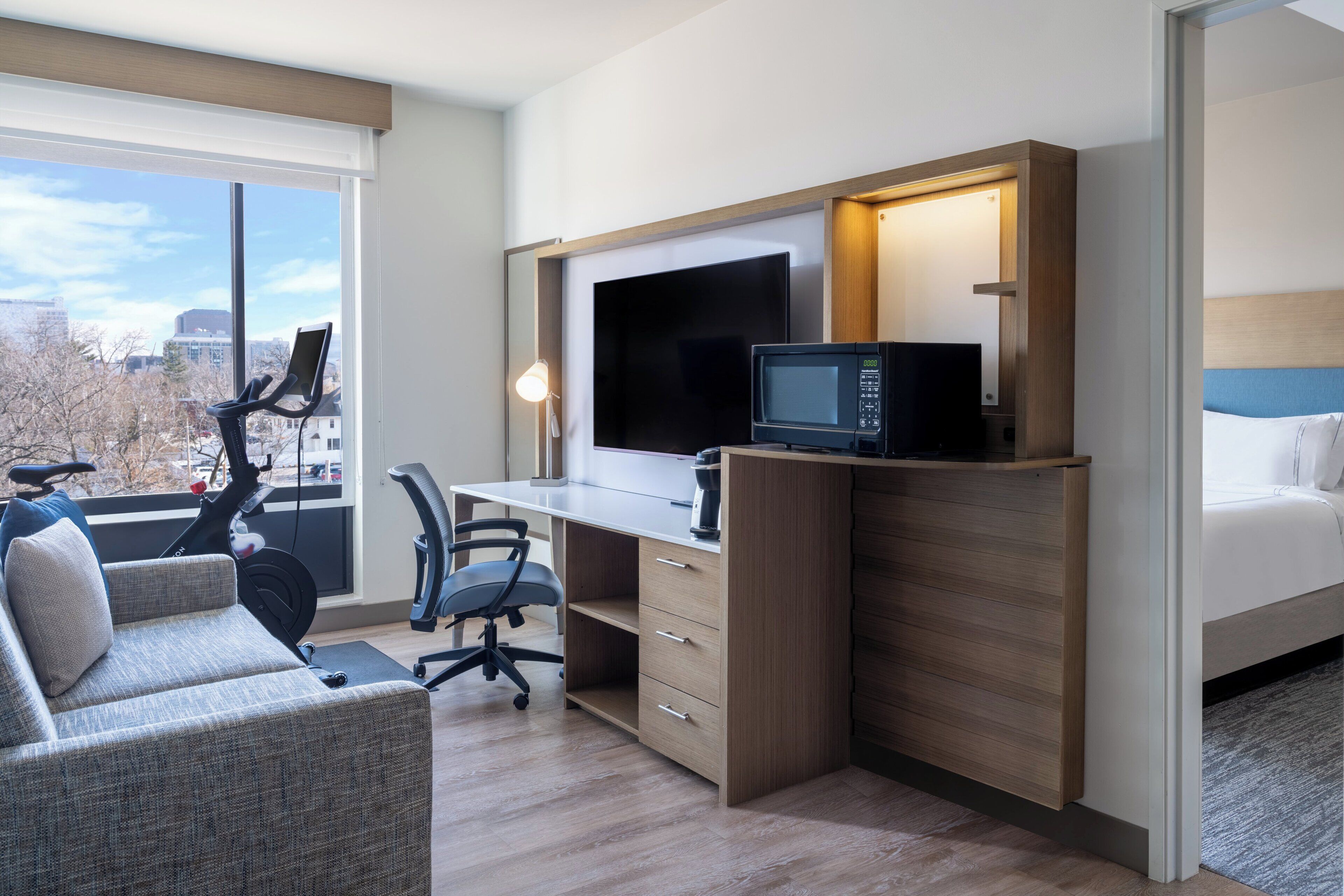 A hotel room with a desk, a chair, a bed, a treadmill, and a large window offering a city view.