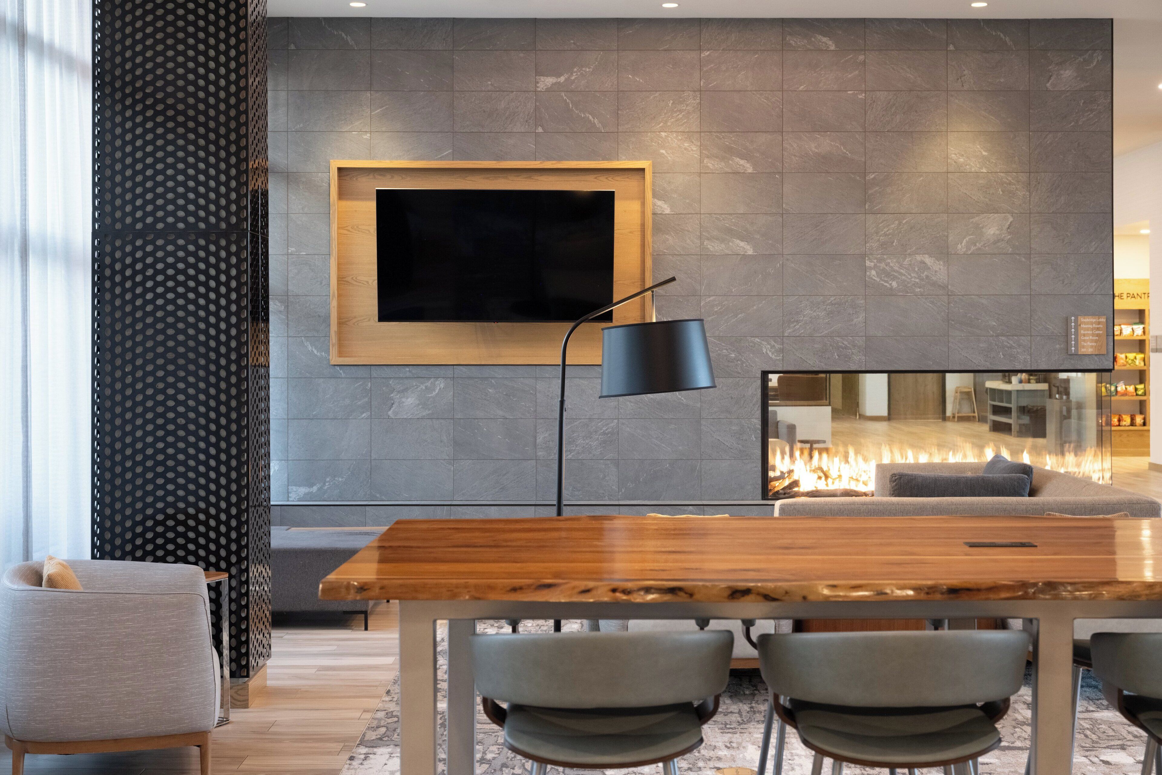 A modern living room with a wooden dining table, grey chairs, a black floor lamp, and a large TV mounted on a grey stone wall.
