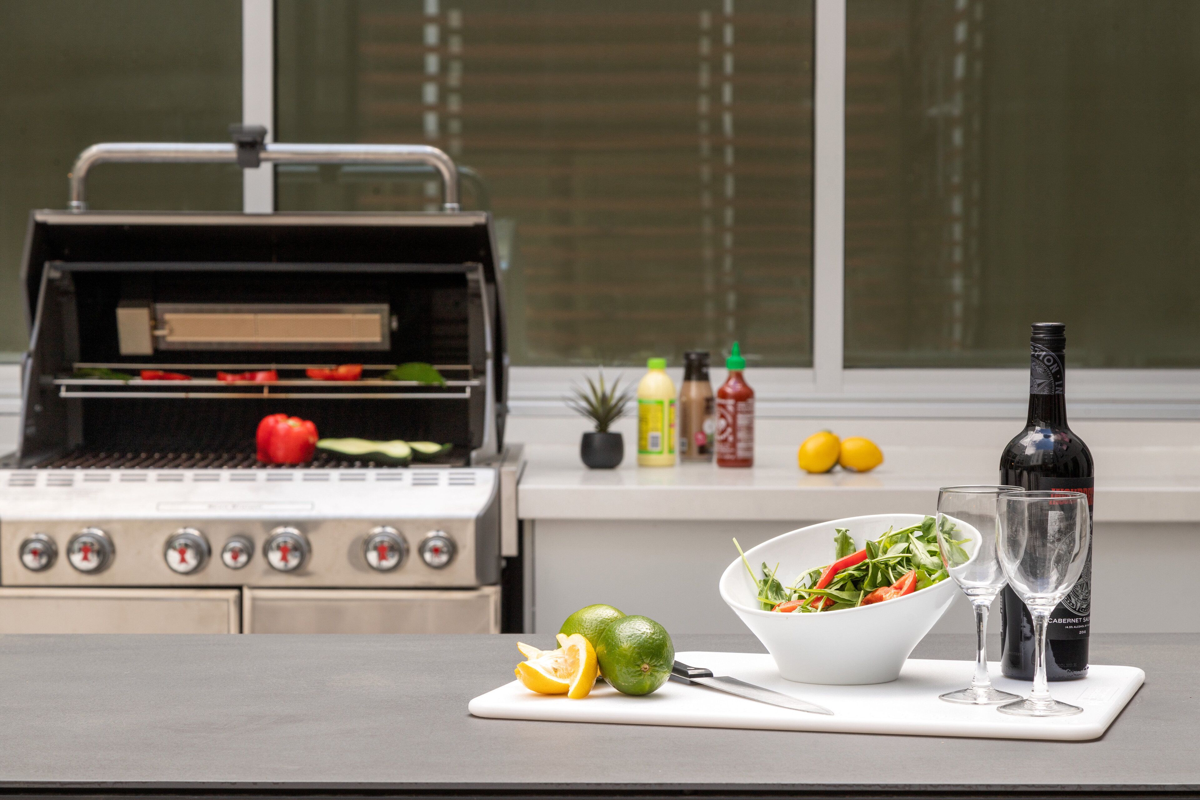 A kitchen with a grill, a cutting board with a lime and a knife, a bowl of salad, two glasses, and a bottle of wine.