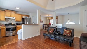 A spacious living area with a black leather sofa, wooden flooring, a kitchen with stainless steel appliances, and a dining area with a chandelier.
