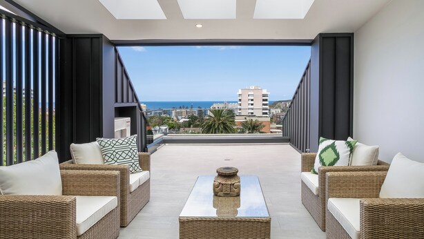 A balcony with wicker furniture and a central table, overlooking a cityscape and the sea. - Designer Penthouse Overlooking Manly, the Ocean and Harbour (Manly)