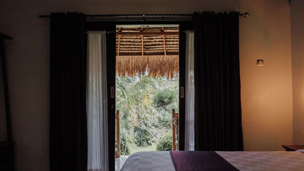 Quarto com cama, janela com cortinas e vista para vegetação lá fora.