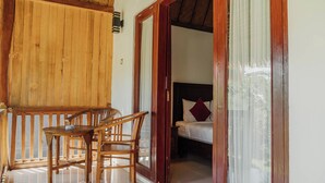 Uma varanda de madeira com mesa e cadeiras, uma cama com travesseiro branco e vermelho, e uma porta de madeira que dá para um quarto.