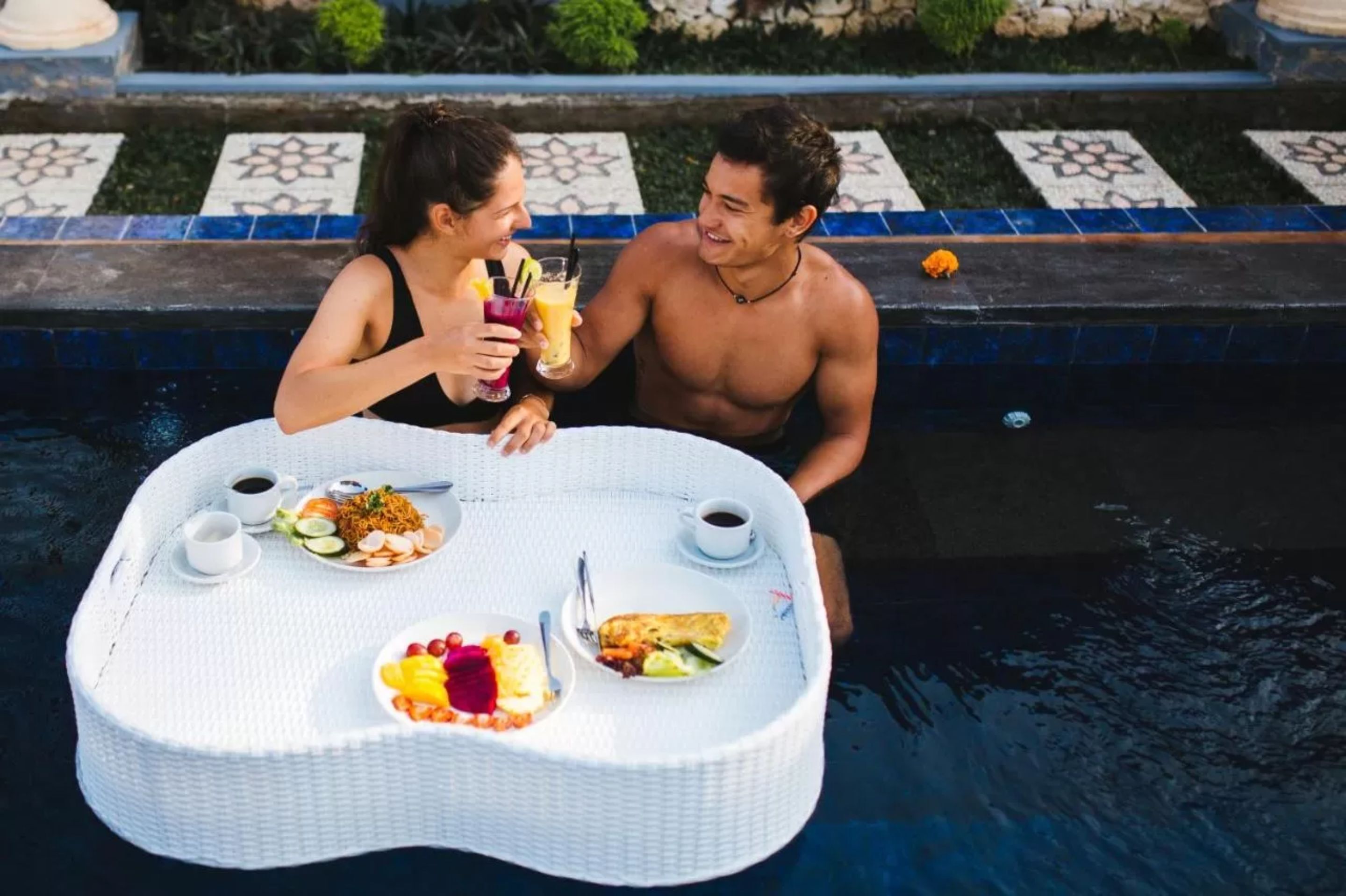 Deux personnes sont assises autour d’une table ronde près d’une piscine, avec de la nourriture et des boissons.