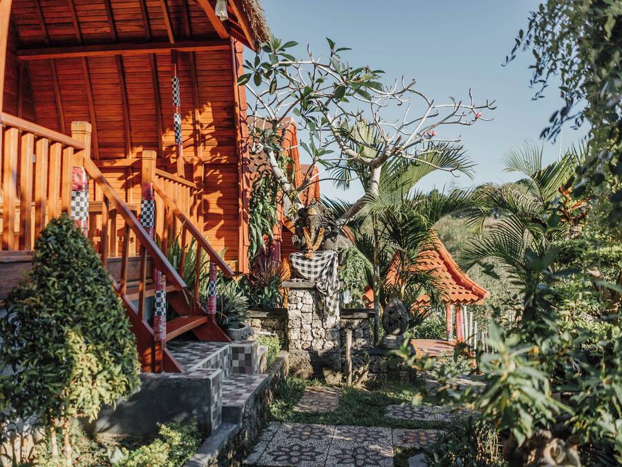 Un edificio de madera con una escalera que lleva a un porche, rodeado de plantas tropicales y un muro de piedra.