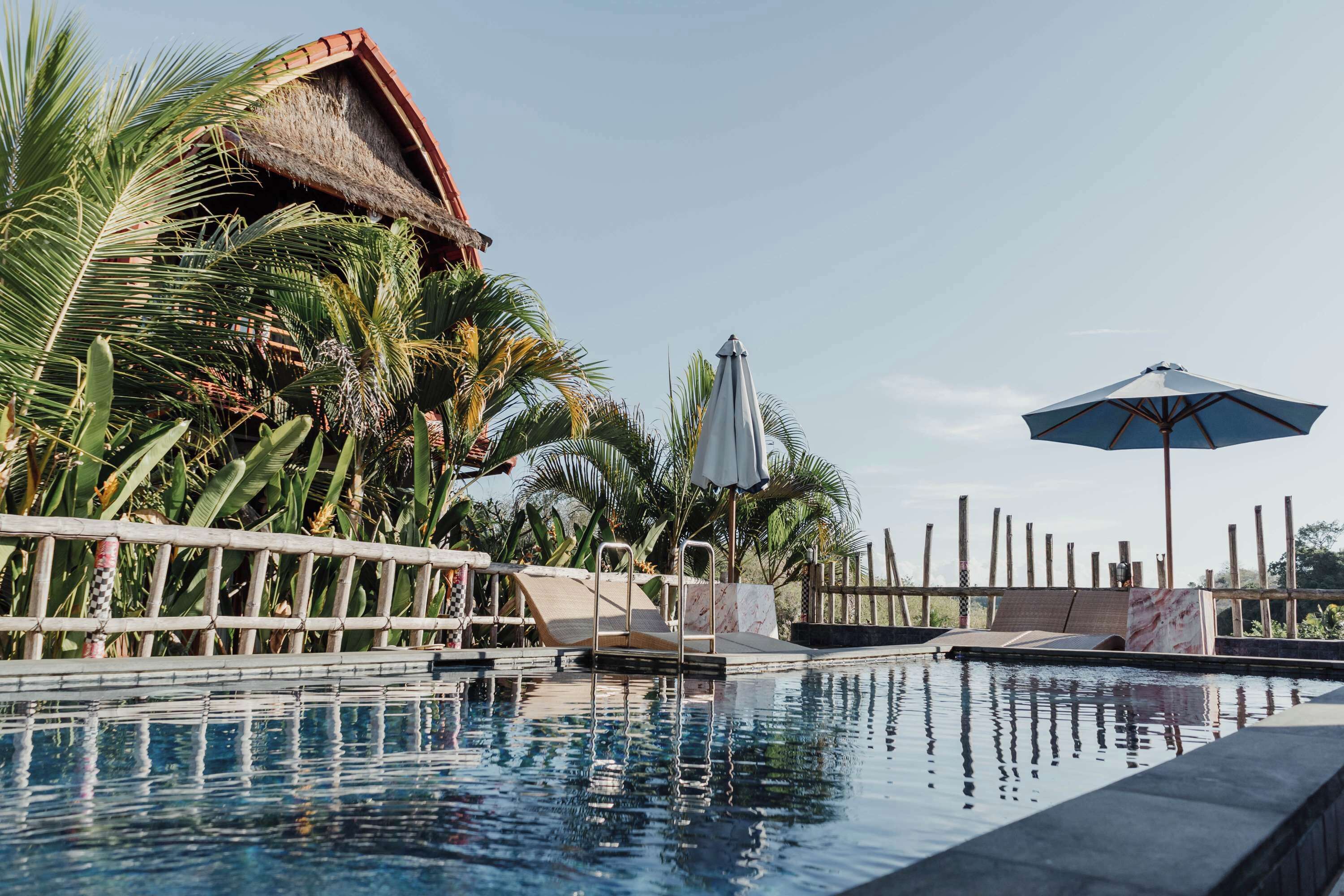 A swimming pool with a thatched-roof structure, palm trees, and lounge chairs.