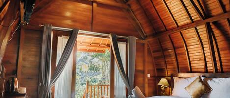 A wooden cabin room with a bed, curtains, a nightstand, and a view of the outdoors.
