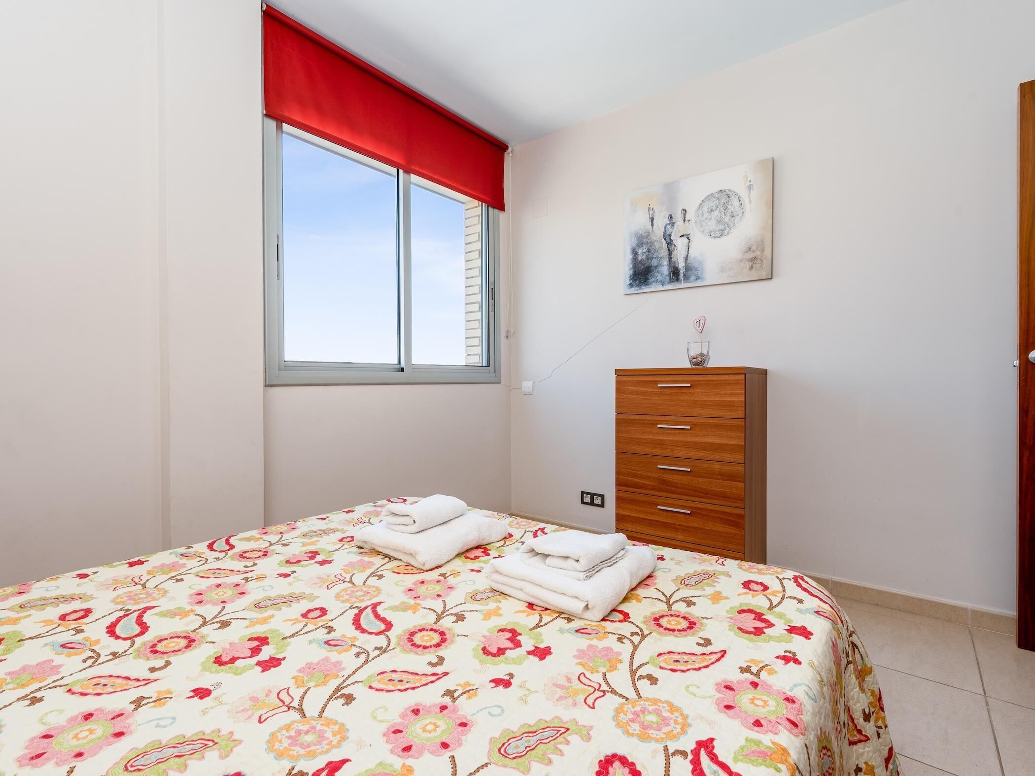 Ein Schlafzimmer mit einem Bett, einem Schminktisch, einem Fenster mit rotem Vorhang und einem Bild an der Wand.