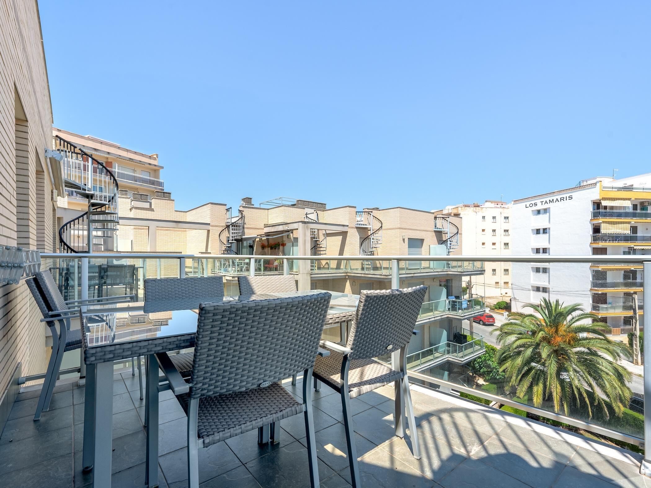 Un balcon agrémenté de fauteuils en osier et d’une table en verre, donnant sur un quartier résidentiel composé de plusieurs immeubles et orné d’un palmier.