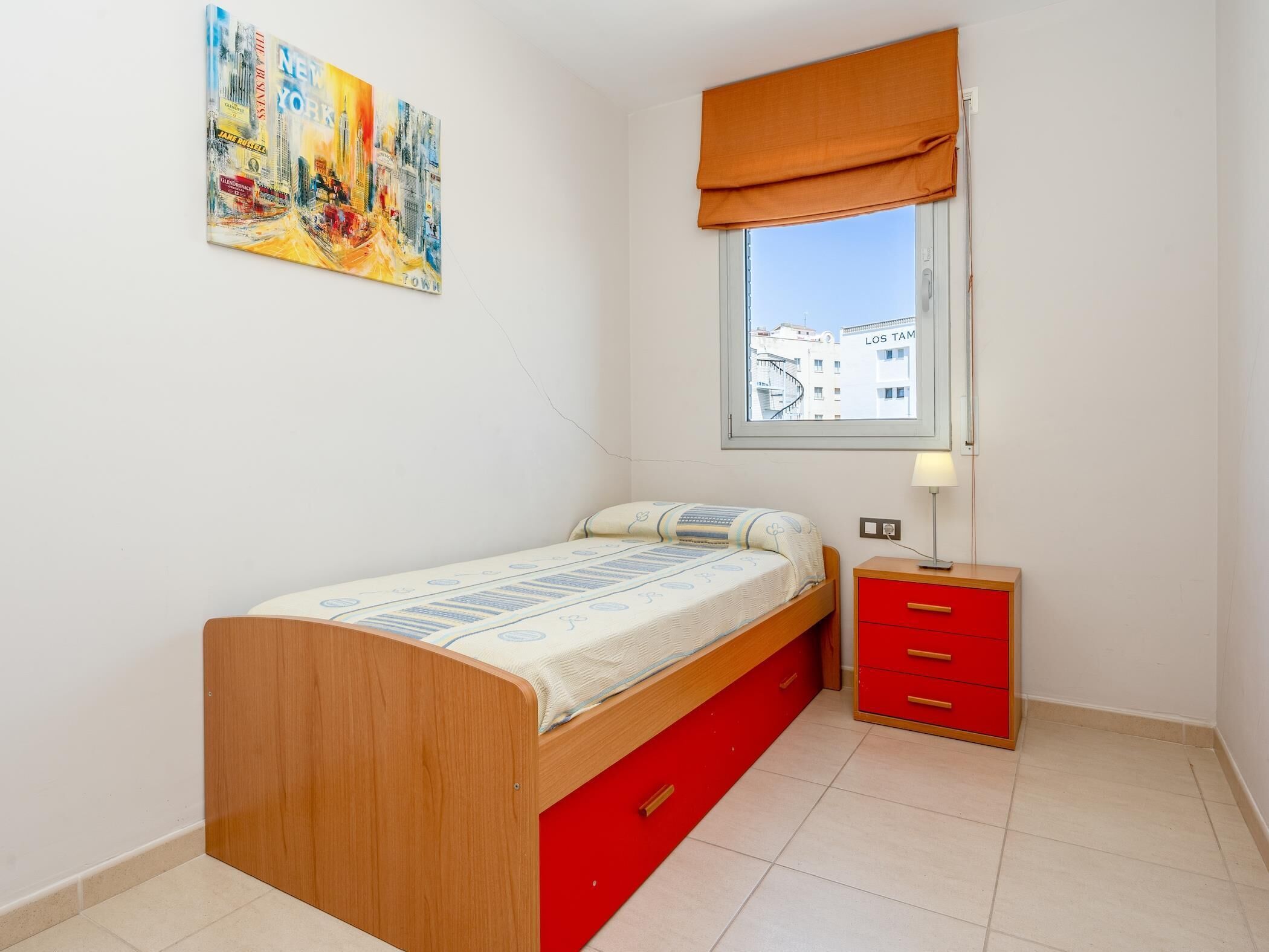 A small bedroom with a wooden bed, a red nightstand, a window with a view of buildings, and a colorful painting on the wall.