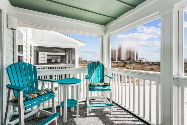 Une véranda couverte avec des chaises de couleur turquoise et une petite table, donnant sur une plage avec des bâtiments visibles au loin.