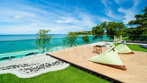 A wooden deck with lounge chairs and a small table, overlooking a clear blue sea and a grassy area with trees. - Charcoal Panwa Beach (Wichit)