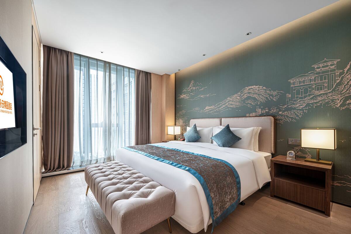 Ein modernes Hotelzimmer mit einem großen Bett, einer Bank, einem Nachttisch mit Lampe und einer Wand mit Wandgemälde.