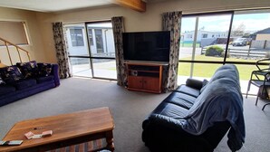 A living room with a sofa, a coffee table, and a television on a stand. There is a large window with curtains, and a view of the outside. - Private holiday home (Twizel)