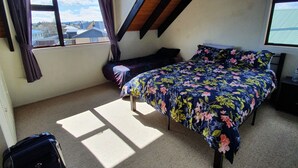 A bedroom with a sloped ceiling, a bed with floral bedding, a suitcase, and a view of buildings through a window. - Private holiday home (Twizel)