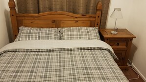 Una cama con colcha y almohadas a cuadros, un cabecero de madera, una mesita de noche con lámpara y un aparador con cajones.