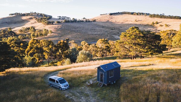 A van parked in a grassy field with a small blue structure nearby. - Deep Creek Sunset by Tiny Away (Monegeetta)