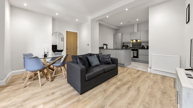 A modern living room with a grey sofa, a dining table with chairs, and a kitchen area with appliances. - Brand New Unique 2 Bedroom Apartment (Reading)
