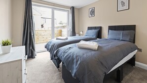 A bedroom with two beds, a sliding glass door to a balcony, a small bedside table, and a potted plant. - Brand New Unique 2 Bedroom Apartment (Reading)