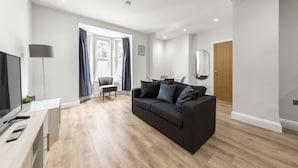 A modern living room with a dark grey sofa, wooden flooring, a TV, and a dining area with a chair and table. - Brand New Unique 2 Bedroom Apartment (Reading)