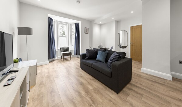 A modern living room with a dark grey sofa, wooden flooring, a TV, and a dining area with a chair and table.