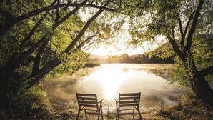 Two wooden chairs facing a calm lake with trees in the background.
