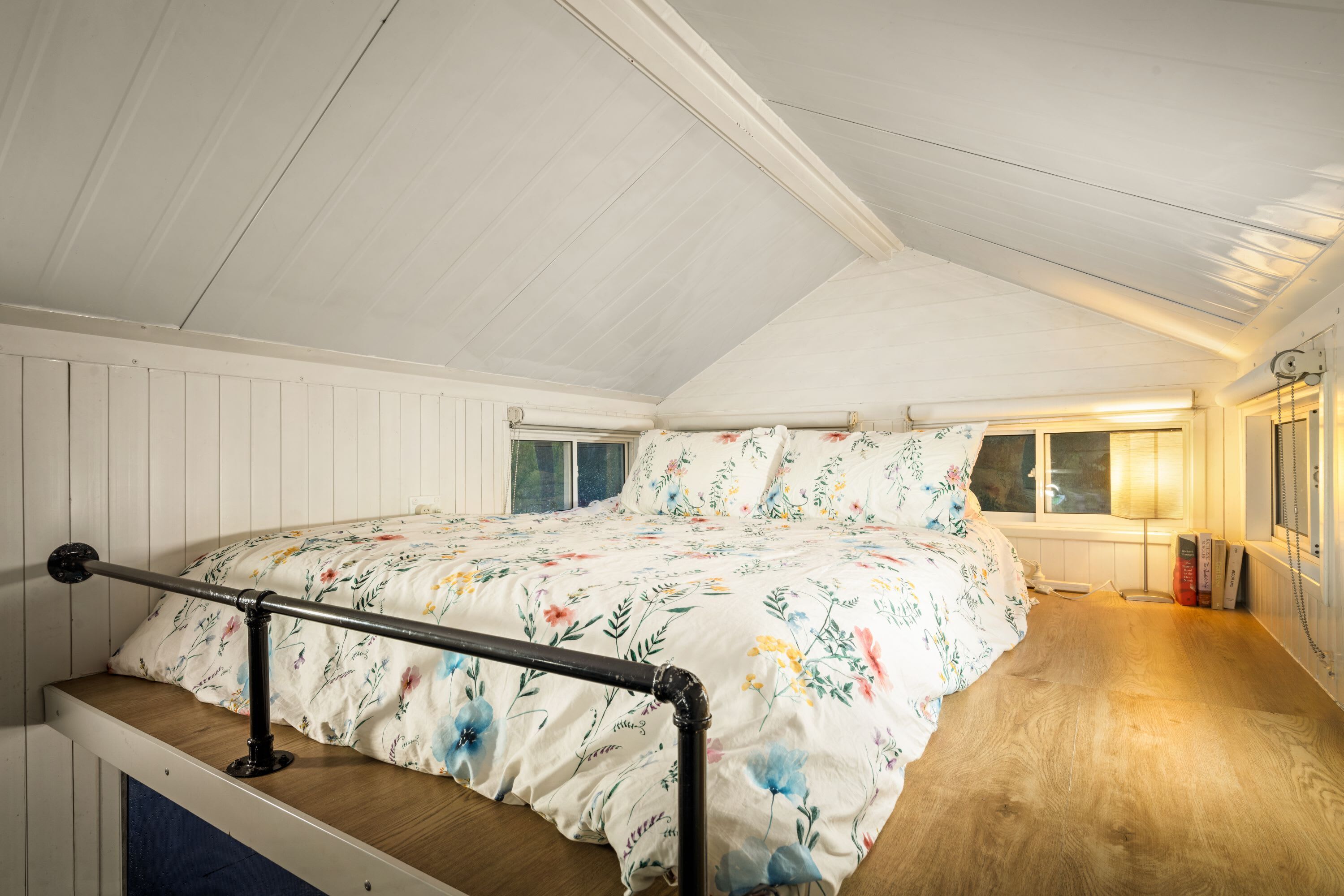 Ein Schlafzimmer mit Holzboden, einem Bett mit Blumendekor, einem Fenster mit Aussicht und einer Lampe auf dem Nachttisch.