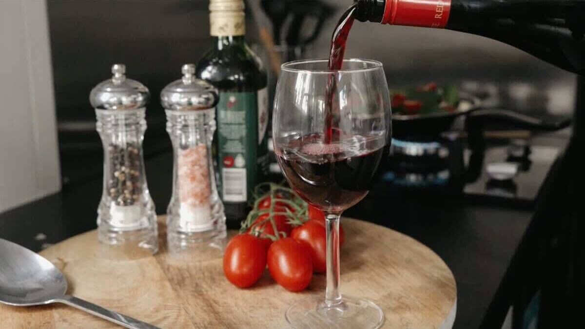 Un verre à vin est en train d’être rempli de vin rouge, entouré de salières et de poivrières, d’une bouteille d’huile d’olive et de tomates fraîches, le tout posé sur une table en bois.