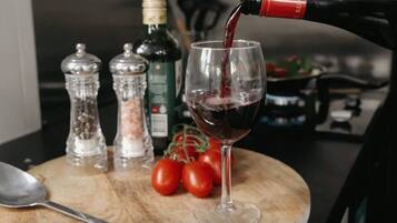 Un verre à vin est en train d’être rempli de vin rouge, entouré de salières et de poivrières, d’une bouteille d’huile d’olive et de tomates fraîches, le tout posé sur une table en bois.