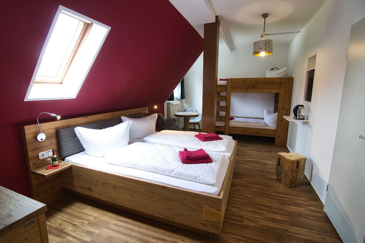 A room with a wooden bed, a small table, a chair, a bunk bed, and a window.