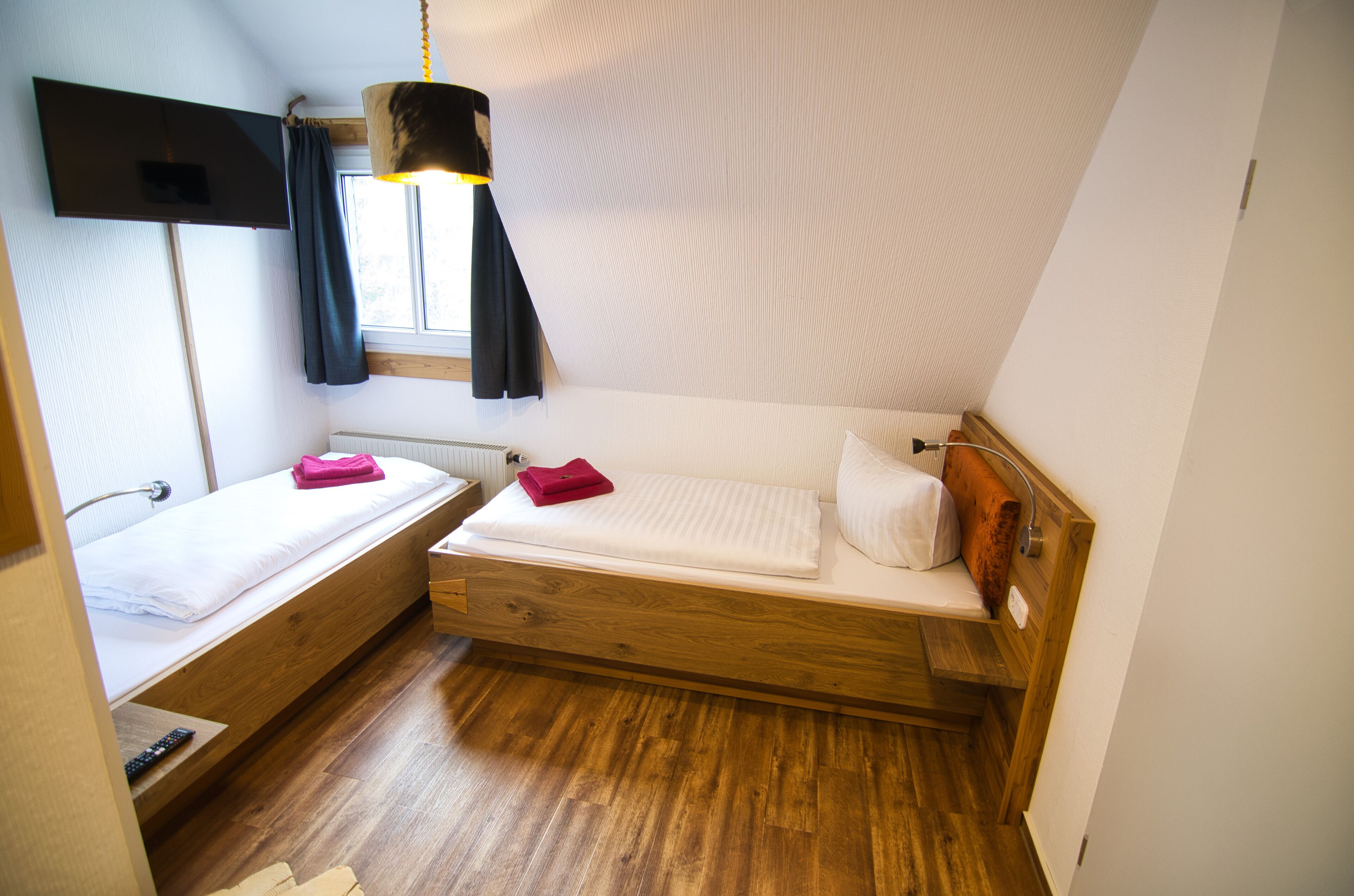A small, compact room with a bed, a bedside table, a TV mounted on the wall, and a window with curtains.
