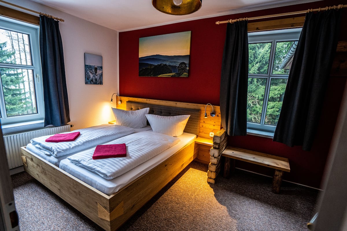 A double bed with a wooden headboard, two pillows, and a red towel. There is a bench and a window with curtains.