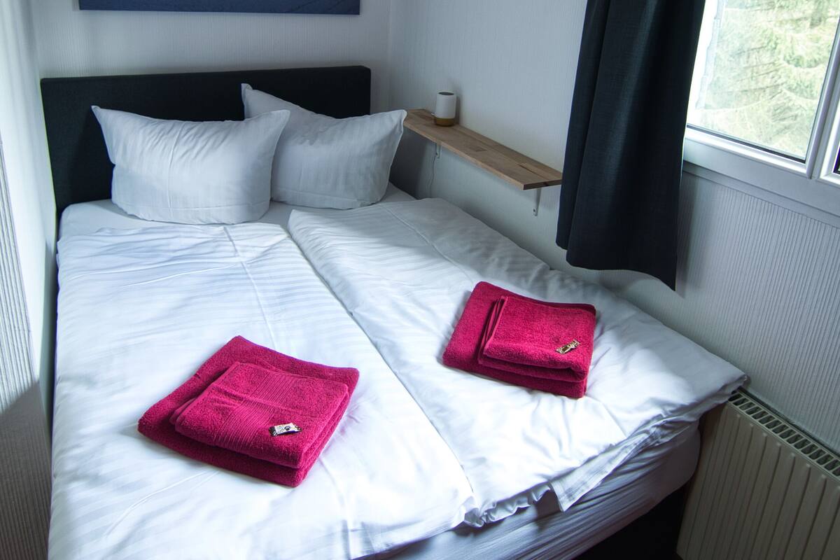 A neatly made bed with white linens and two pink towels, a bedside table with a lamp, a window with dark curtains, and a painting of a sunset on the wall.
