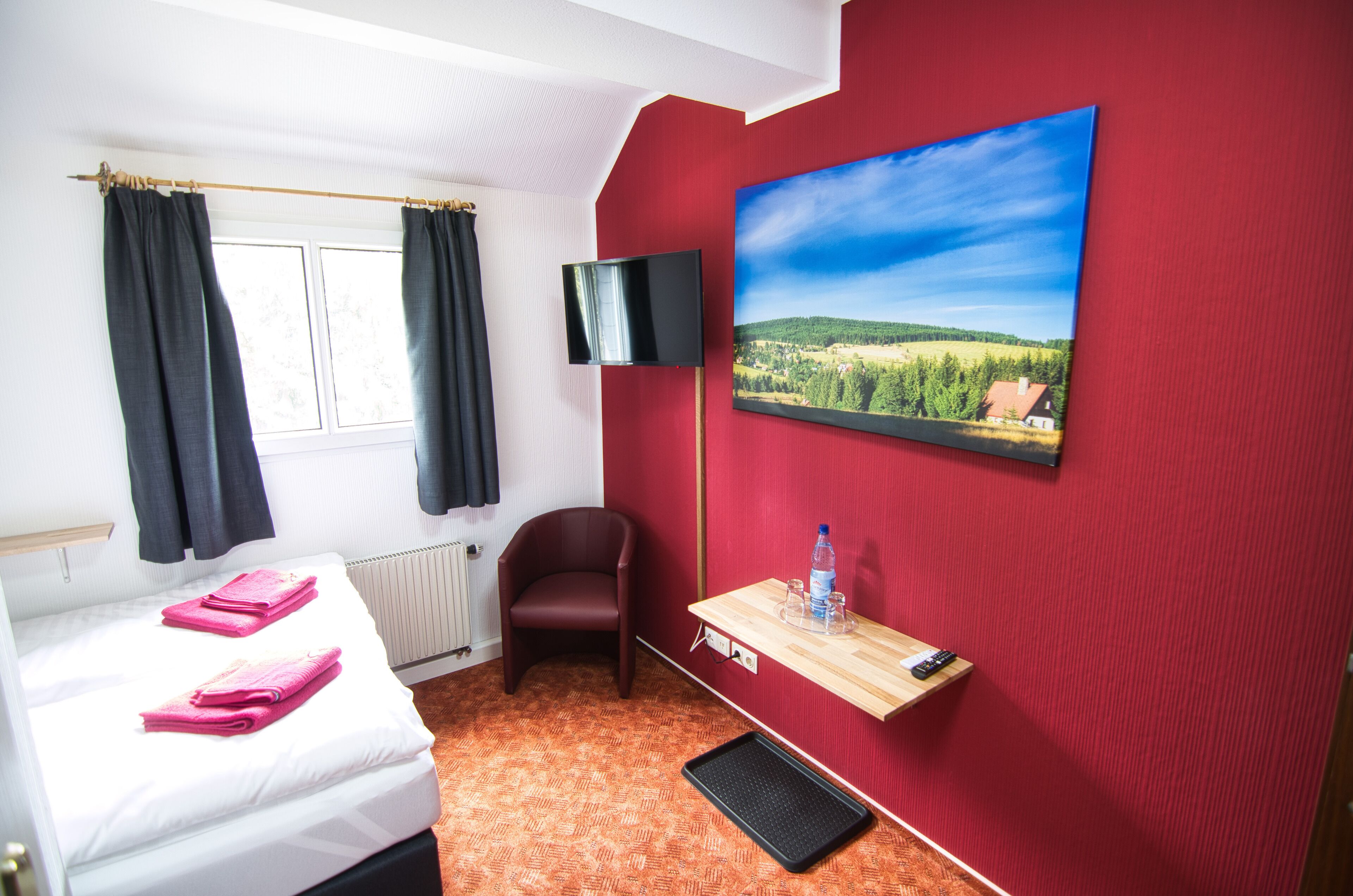 Quarto de hotel com cama, uma cadeira, televisão, pintura e uma pequena mesa com garrafa e copos.