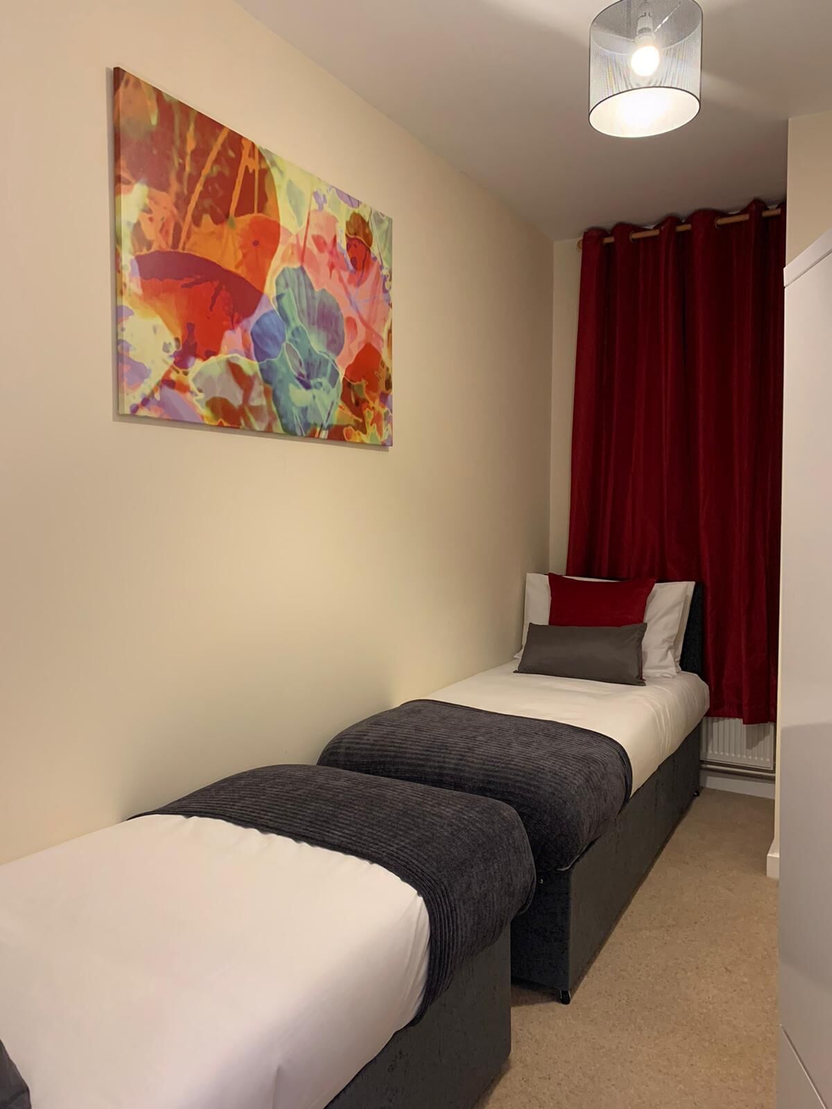 A small, modern room with two beds, a colorful abstract painting, and red curtains.