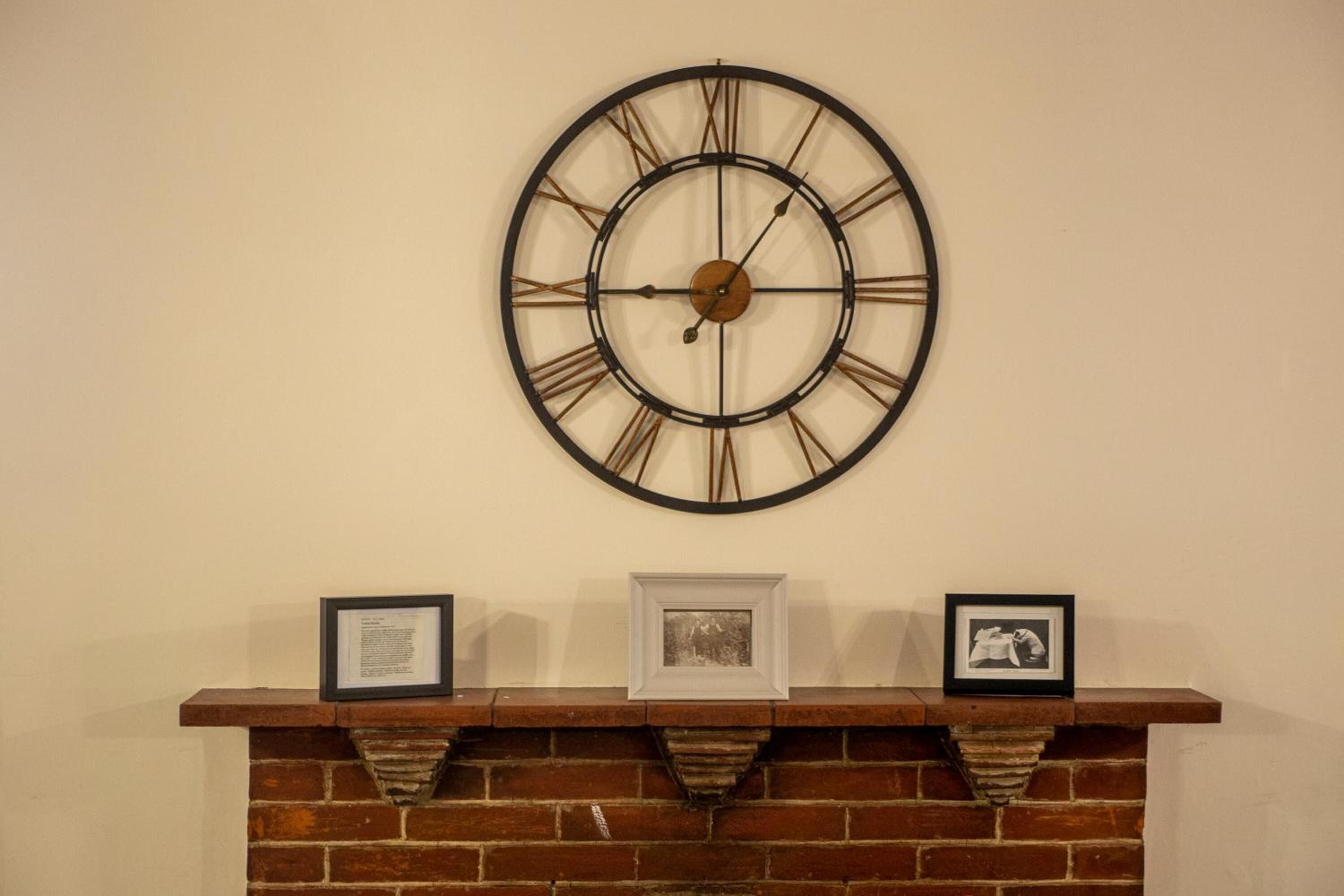 Een wandklok met Romeinse cijfers en een open haard met drie ingelijste foto's.