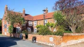 A traditional brick building with a red-tiled roof, multiple chimneys, and a well-maintained garden with various plants and a palm tree.