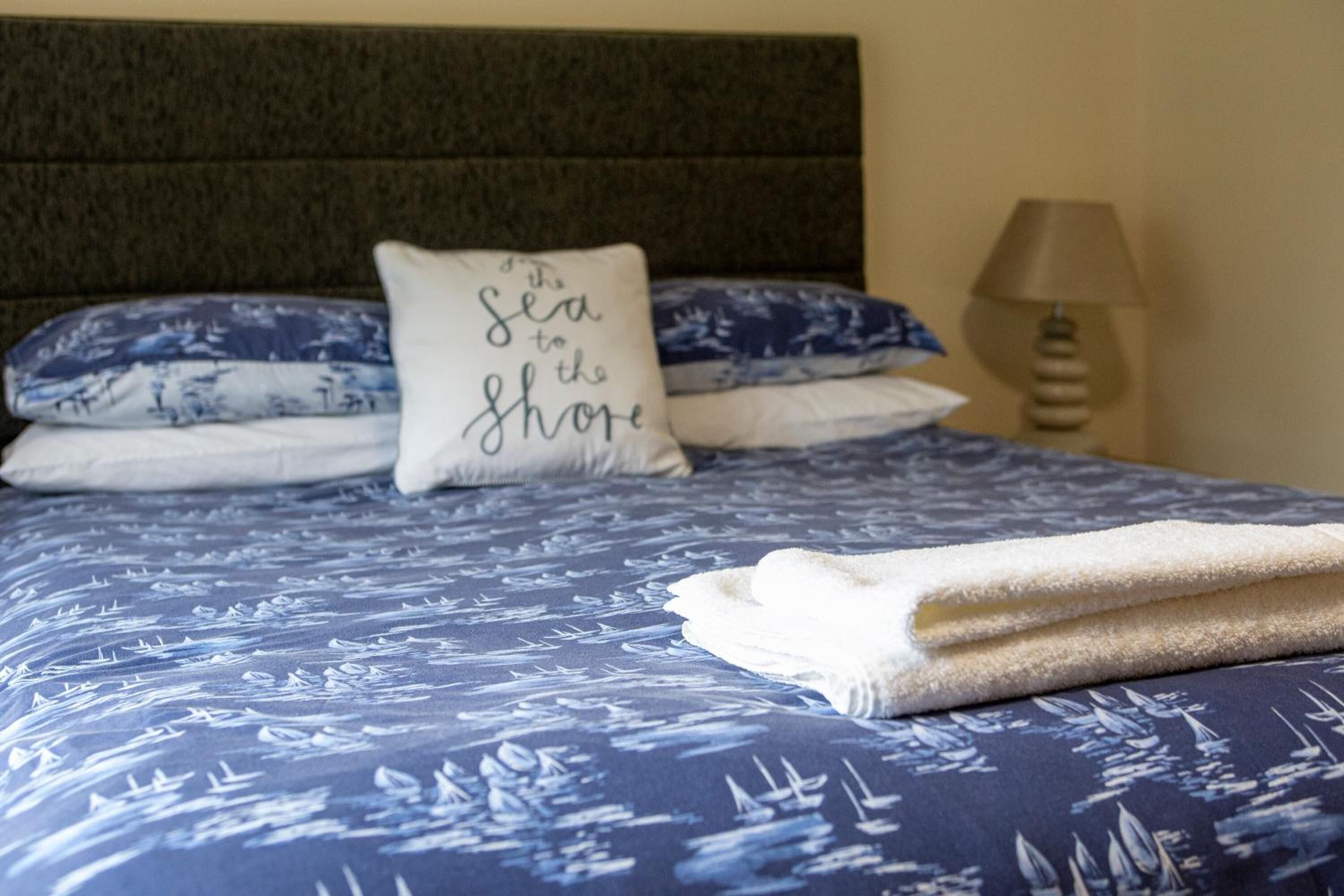 A neatly made bed with a blue and white nautical-themed bedspread, a pillow with the text "the sea to the shore," and a folded white towel on the bed.
