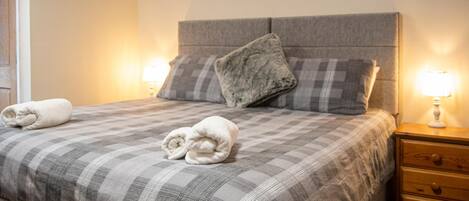 A neatly made bed with a checkered bedspread, two pillows, and two rolled towels. There is a nightstand with a lamp and a drawer.