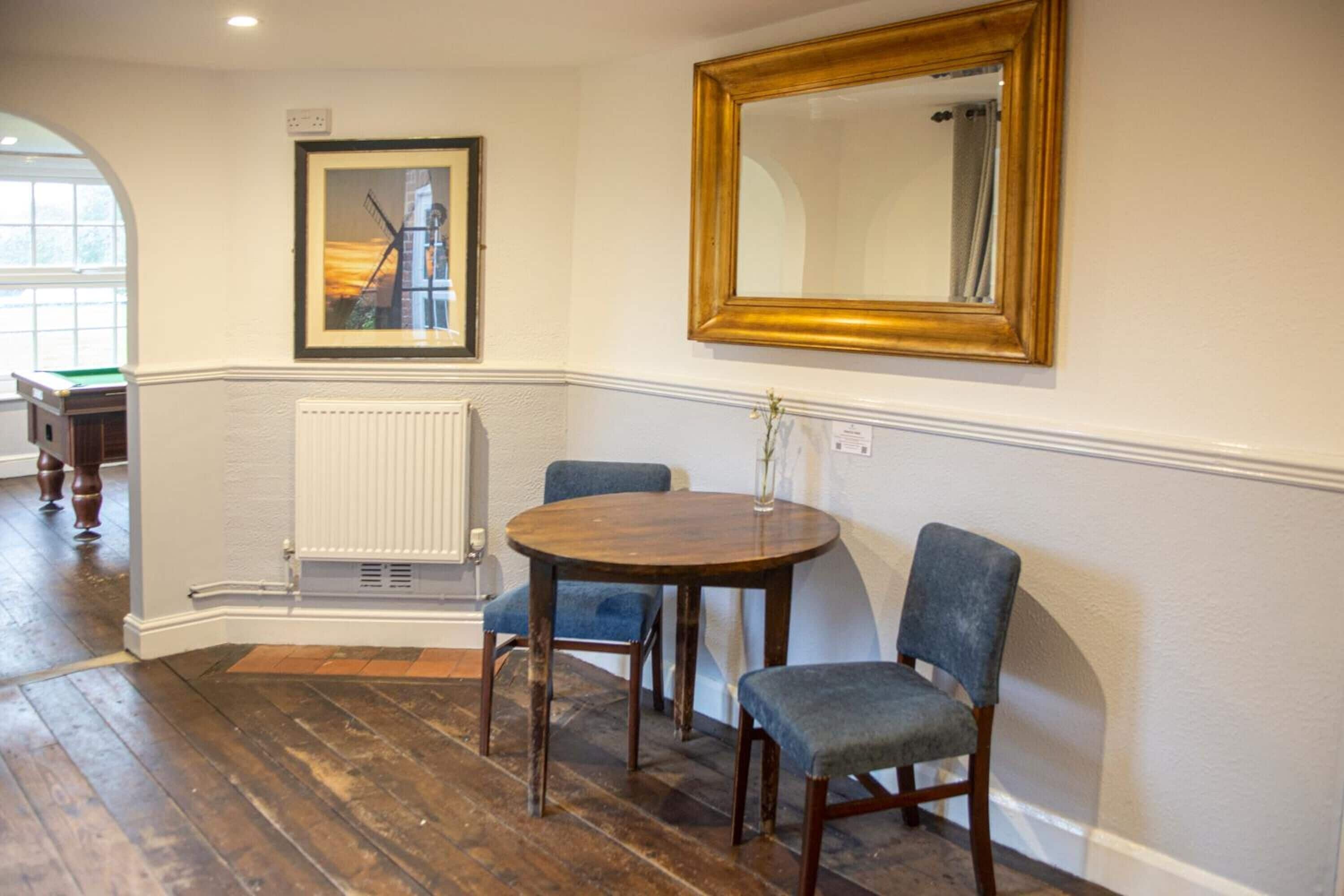 Una habitación con una mesa redonda de madera, dos sillas tapizadas en azul, un cuadro enmarcado y un espejo grande en la pared.