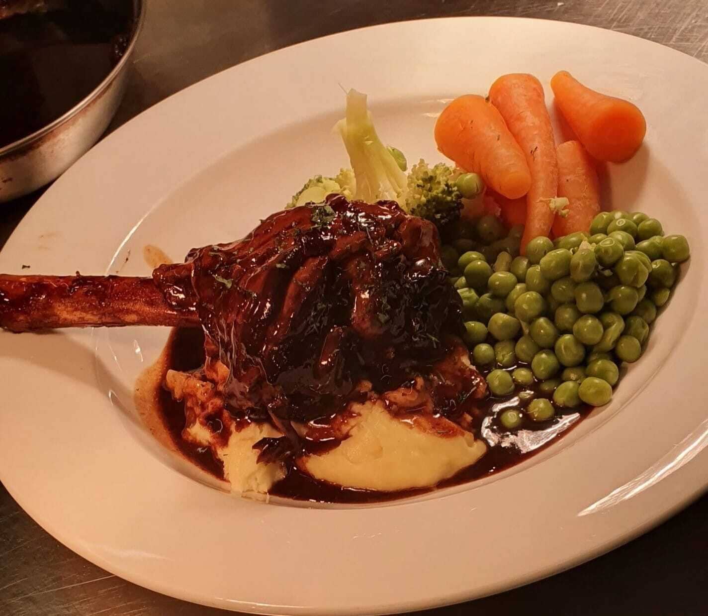 A plate with a lamb shank, mashed potatoes, peas, carrots, and broccoli.