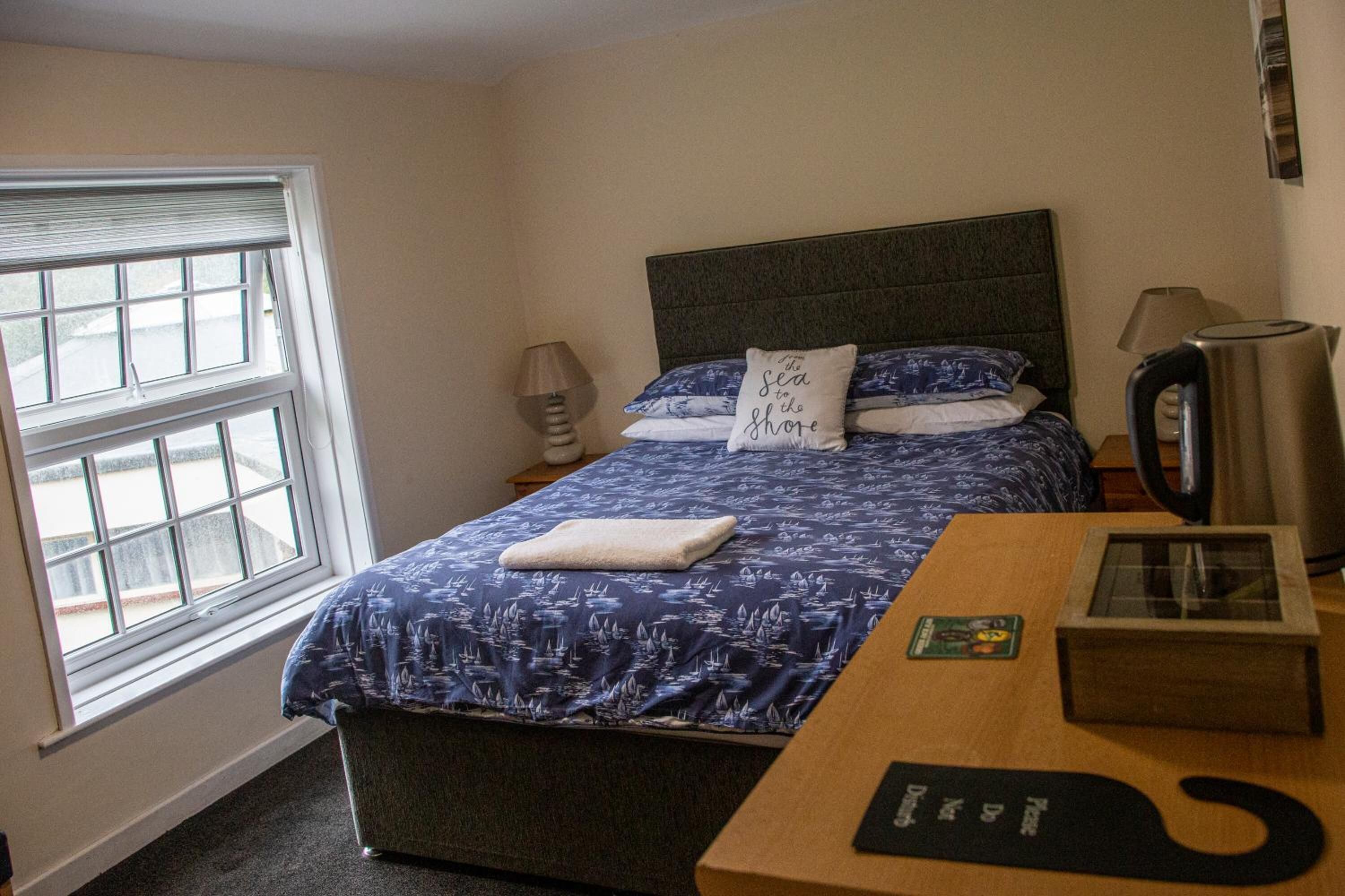 Una habitación de hotel con cama, mesita de noche, lámpara, hervidor de agua y ventana.