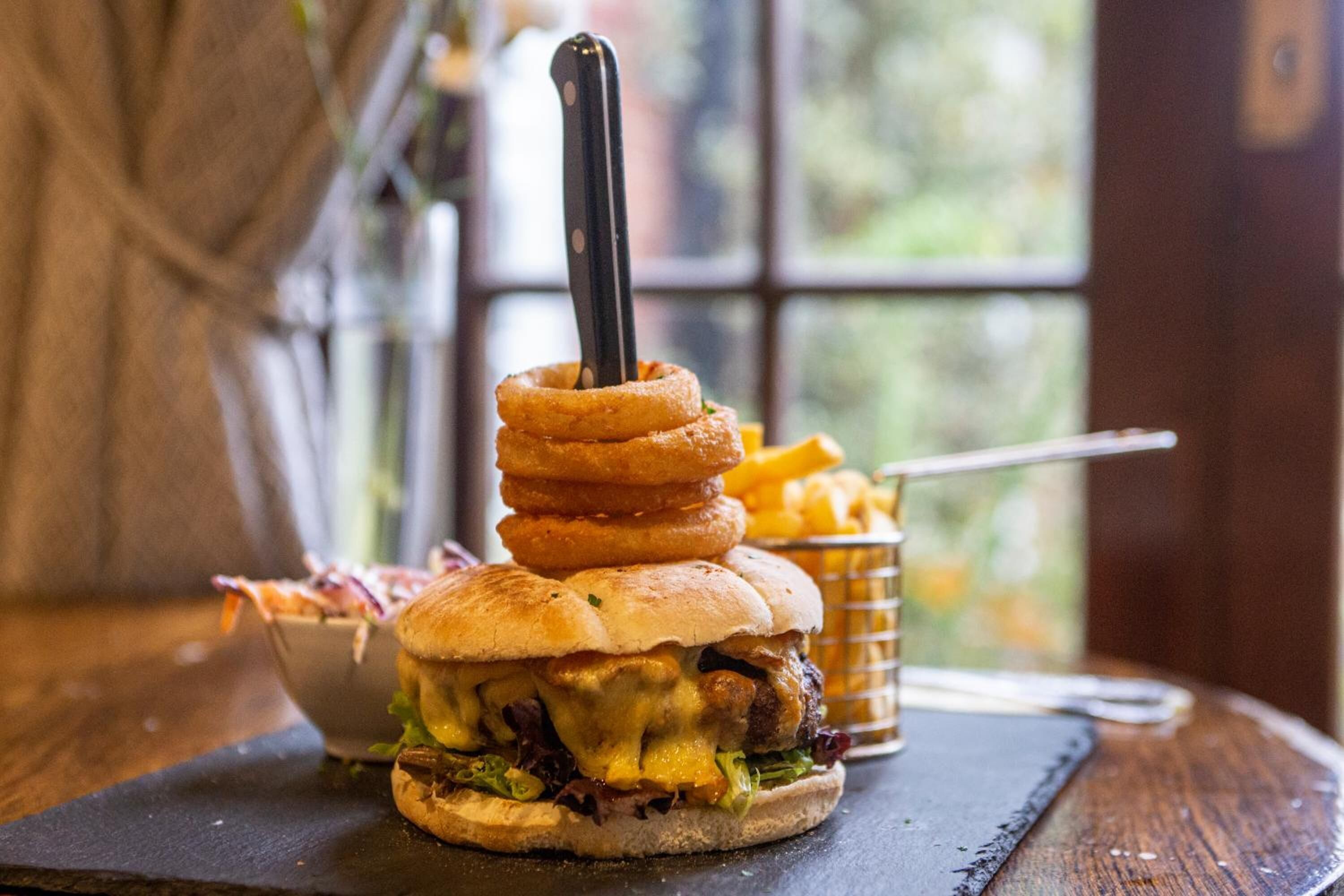 A burger with stacked onion rings and a knife on top, served with fries and a side salad.