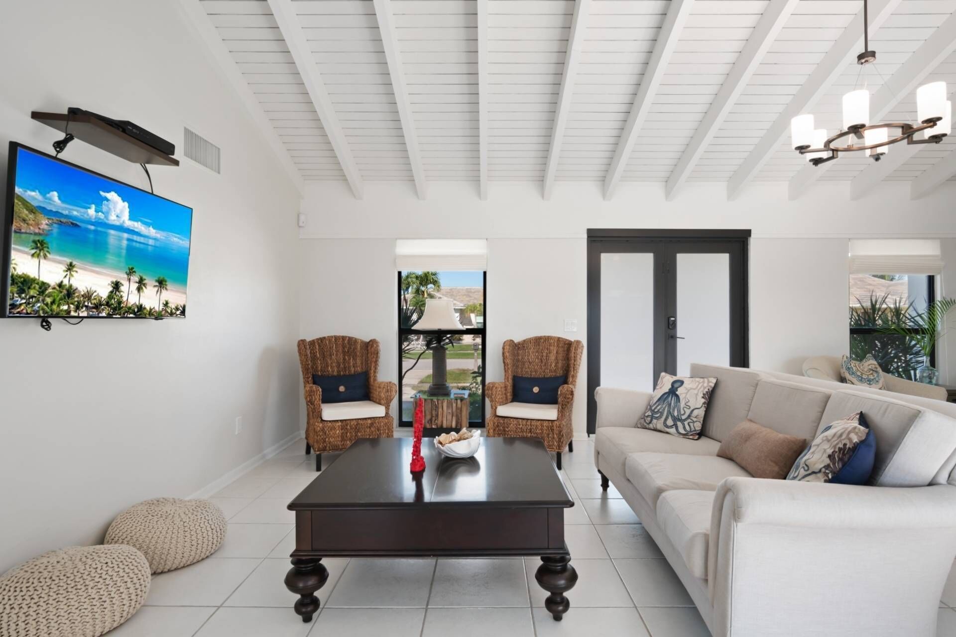 Un salón espacioso con un televisor de pantalla plana grande, dos sillas de mimbre, una mesa de centro de madera oscura y un sofá blanco con muchos cojines.
