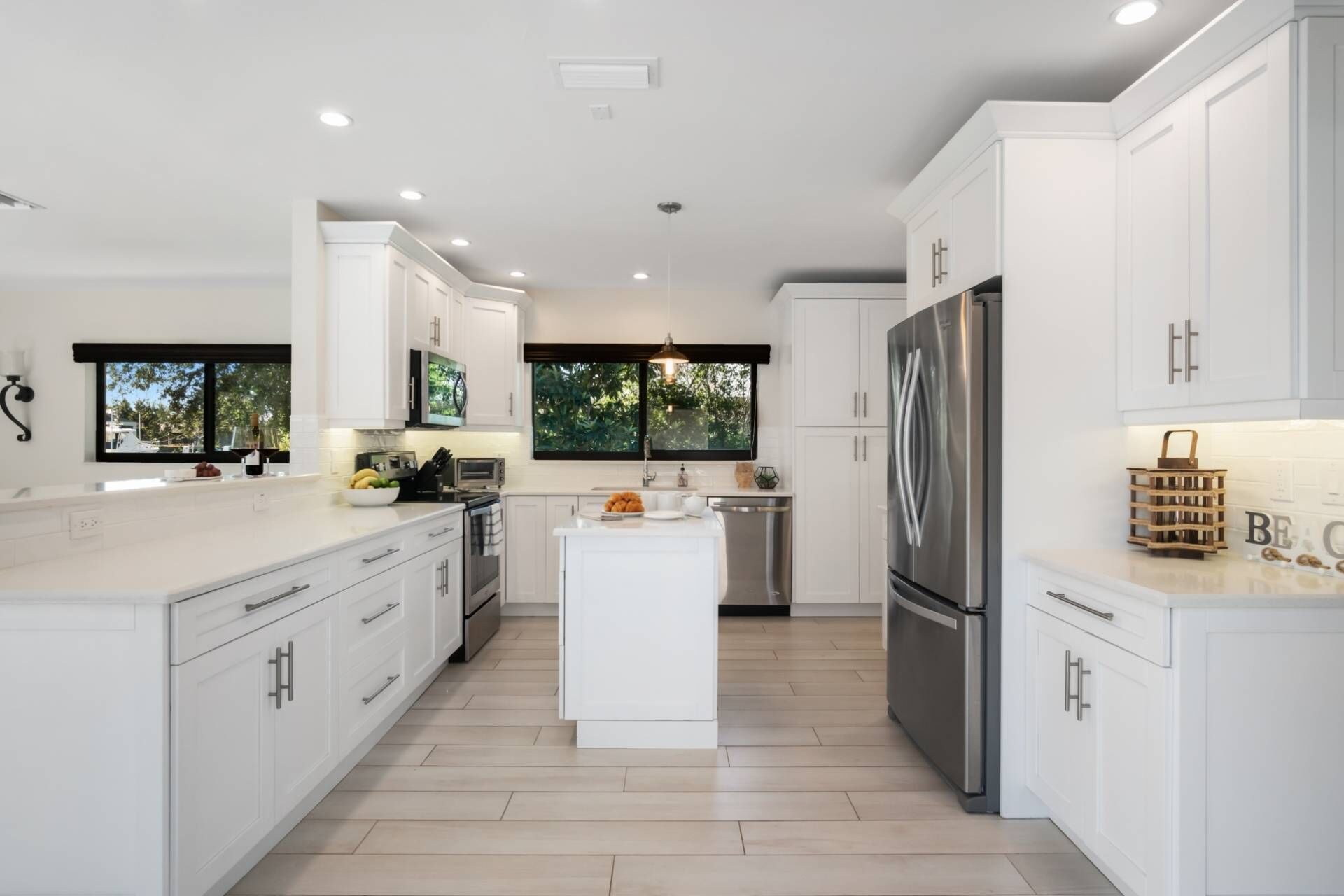 A modern kitchen with white cabinetry, a central island, a stainless steel refrigerator, and a view of greenery outside.