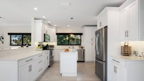 A modern kitchen with white cabinetry, a central island, a stainless steel refrigerator, and a view of greenery outside.